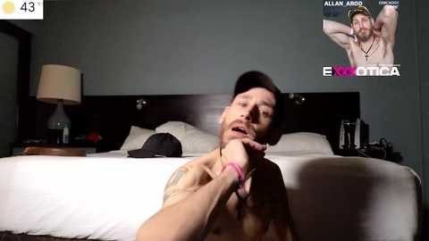 Media: Video of a shirtless, muscular man with tattoos, sitting on a bed, holding a black cap, in a dimly-lit room with a gray wall, a lamp, and a poster of a shirtless man.