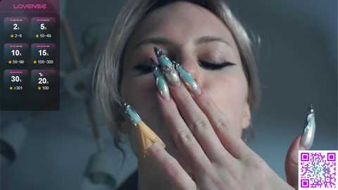 Video of a woman with light skin, blonde hair, and long, elaborate nail art featuring metallic gold and black designs, wearing a black top. The background is blurred, with a \"Live Now\" overlay on the left. QR code in bottom right corner.