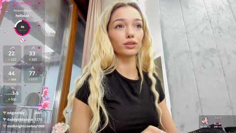 Video of a young woman with long, wavy blonde hair and fair skin, wearing a black T-shirt, standing indoors with a modern, light-colored wall and large window in the background.