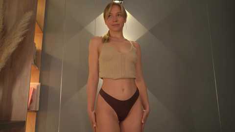 Media: Video of a slim, Caucasian woman with blonde hair in a ponytail, wearing a beige crop top and dark brown panties, standing in a modern, dimly lit room with geometric-patterned walls.