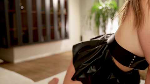 Media: Video of a woman with light skin, wearing a black, shiny, strapless dress, holding a black purse. Background features a modern living room with wooden floors, a potted plant, and a bookshelf.