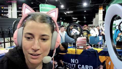 Media: Video of a woman with light skin, brown hair, wearing pink cat ear headphones, smiling, at a crowded convention booth with a large round mirror, a cake, and a 'No Contact Card' sign.