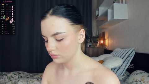 Media: Video of a light-skinned, dark-haired woman with brown eyes, wearing minimal makeup, sitting on a bed with floral sheets, looking down. Background shows a dimly lit room with a lamp and dark curtains.
