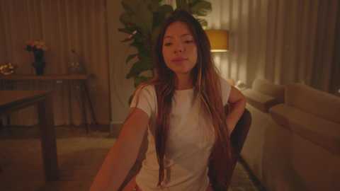 Media: Video of an Asian woman with long, dark hair, wearing a white blouse, sitting on a couch in a warmly lit, cozy living room with beige curtains and a potted plant.