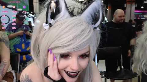 Video of a woman with light skin, wearing a silver wig, cat ears, black lipstick, and purple nail polish, smiling at the camera, in a crowded convention hall with colorful banners and people in the background.