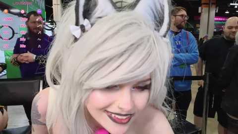 Media: Video of a woman with platinum blonde hair, wearing cat ears, black lipstick, and a pink choker, smiling at an anime convention. Background features a mix of attendees in casual clothes and a large screen displaying Japanese text.