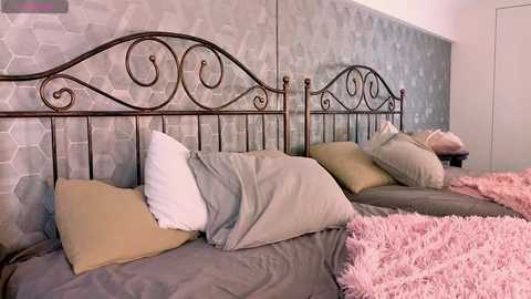 Media: Video of a cozy bedroom featuring two wrought iron beds with floral headboards, covered in light gray and beige bedding, and flanked by a textured gray wall and a fluffy pink throw rug.