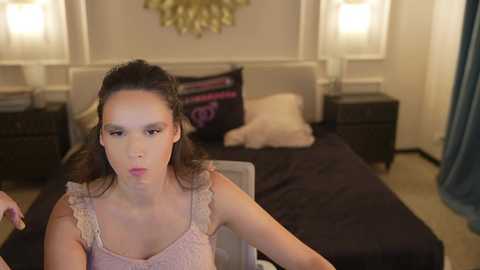 Video of a young woman with fair skin, brown hair, and dark makeup, sitting in a dimly-lit bedroom with a black bed, white walls, and two bedside lamps.