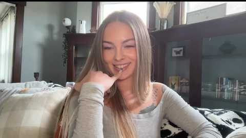 Video of a smiling, long-haired, light-skinned young woman in a gray sweater, holding a sandwich, sitting on a patterned couch in a cozy living room with bookshelves and a window.