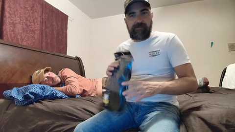 Video of a bearded man in a white t-shirt and blue jeans holding a large, dark-colored dildo in a bedroom with a brown bed, a man in an orange shirt and hat on the left.