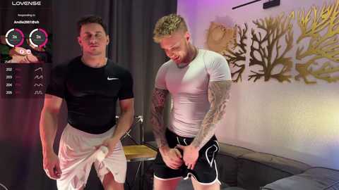 Video of two muscular men in workout gear, one adjusting his shorts, in a room with a grey couch, coral painting, and digital stats.