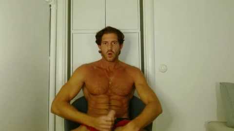 Video of a shirtless, muscular, tan-skinned man with short brown hair, sitting in a chair, giving a thumbs-up gesture. He wears red shorts. Background shows a white door and wall.