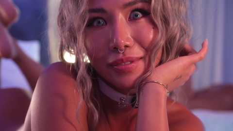 Video of a young woman with light brown, wavy hair and light skin, wearing a nose ring, smiling with her hands supporting her chin. Background is blurred with purple and white tones.