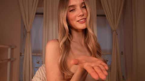 Video of a fair-skinned, blonde woman with long hair, smiling, extending her hand, wearing a beige dress. Background features beige curtains and a window.