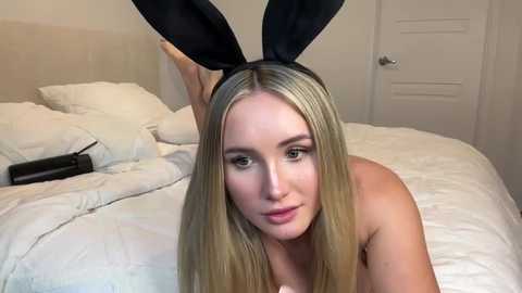 Video of a blonde woman with fair skin, wearing black bunny ears, lying on a bed with white sheets.
