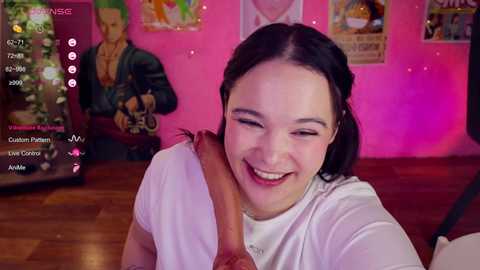 Video of a smiling young woman with dark hair and pale skin, wearing a white t-shirt, leaning in to kiss a large erect penis. Background features a pink wall, posters, and a green statue.
