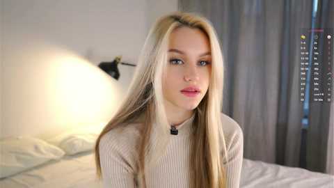 Media: Video of a young blonde woman with long, straight hair, wearing a beige ribbed sweater, sitting on a bed in a softly lit bedroom with white curtains and a black wall lamp.