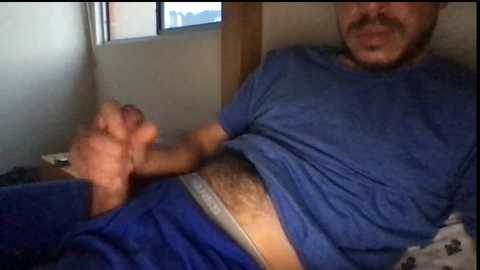 Video of a man with a beard, wearing a blue shirt and blue shorts, holding his erect penis in a dimly lit room.