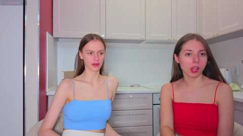 Video of two young women with long brown hair, one in a blue tank top and the other in a red tank top, sitting in a modern kitchen with white cabinets and light countertops.