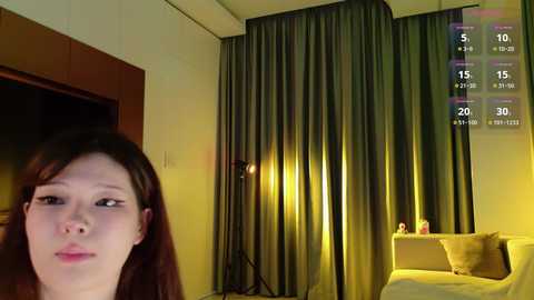 A video of a modern bedroom with a woman in the foreground, brown hair, wearing a white top. Background features dark curtains, a lamp, and a TV screen showing a weather forecast.