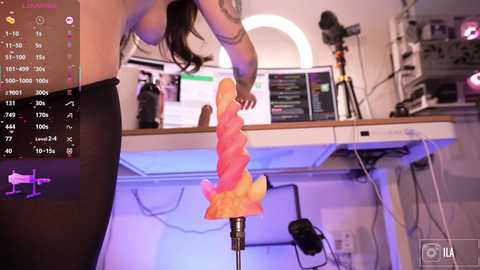 Video of a woman in a purple bra, holding a pink, phallic sex toy in a modern, dimly lit room with a desk, monitor, and microphone.