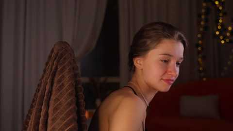 Video of a young woman with light skin, brown hair, and a thin build, wearing a black strapless top, standing against a dimly lit, warmly colored bedroom background.