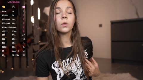 Media: A young, light-skinned girl with long brown hair, wearing a black graphic t-shirt, closes her eyes and leans forward, in a dimly lit room. A screen displays video stats.
