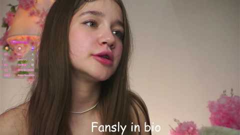 Video of a young, fair-skinned girl with long brown hair, wearing a silver necklace, looking introspective against a pink and white background. Text: \"Fansly in bio.\