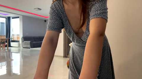 Video of a woman with light brown skin, medium build, and brown hair, wearing a low-cut, black and white floral dress, bending over in a modern, minimalist living room with a sleek gray sofa, glass coffee table, and red LED strip lighting.