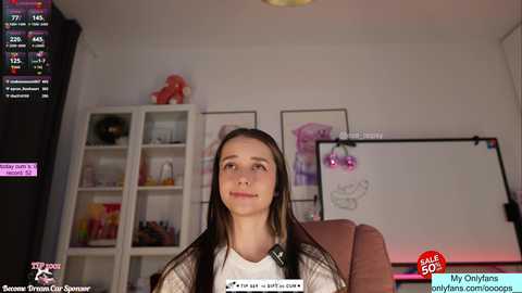 A video of a young woman with long brown hair and fair skin, wearing a plaid shirt, sitting in a modern room with a projector screen and white shelving unit.