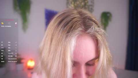 Media: Video of a blonde woman with shoulder-length hair, blurred background featuring green plants and a calendar.