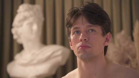 Video of a shirtless, pale-skinned young man with short dark hair, looking contemplative, standing against a blurred background featuring a white bust sculpture and wooden panels.