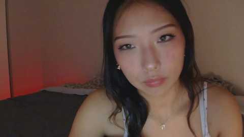Video of an East Asian woman with long black hair, fair skin, wearing a light gray tank top, and small gold earrings, indoors with dim lighting.