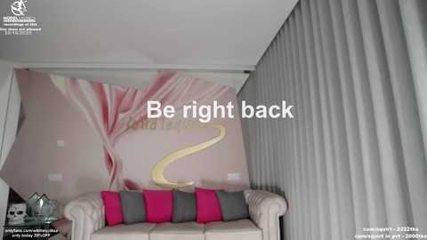 Video of a modern living room featuring a pink and white floral wallpaper, a beige couch with pink and gray throw pillows, and a cream-colored lamp. Text reads, \"Be right back.\