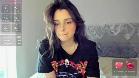 A video of a young woman with long, dark hair, wearing a black T-shirt with a colorful graphic design. She stands in a softly lit bedroom with white lace curtains and a bed with grey bedding.