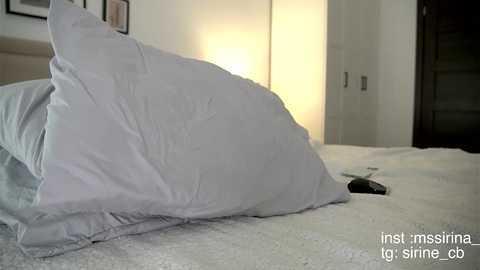 Media: Video of a neatly made bed with white pillows and a white duvet, featuring a minimalist, modern bedroom with a beige headboard and a soft, ambient light.