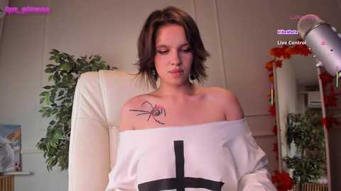 Video of a fair-skinned woman with shoulder-length brown hair, wearing an off-shoulder white dress, featuring a black cross and a spider tattoo. She's indoors, surrounded by greenery and a mirror.