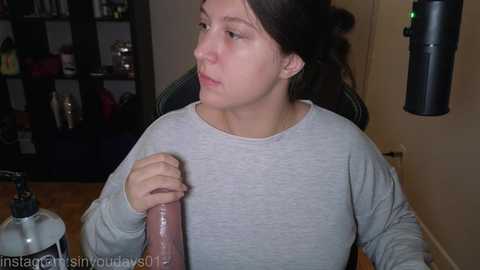A video of a woman with fair skin and dark hair in a ponytail, wearing a light gray sweater, holding a large, veiny, pink dildo. The room has shelves filled with bottles and a microphone stand in the background.