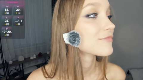 A video of a young woman with fair skin, straight brown hair, and a fluffy grey makeup sponge on her cheek, taken in a modern, minimalistic bedroom.