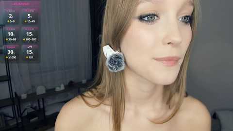 Video of a fair-skinned, light-haired woman with blue eyeshadow, wearing a black headband and a fluffy makeup brush on her ear, in a modern indoor setting with a virtual makeup counter display in the background.