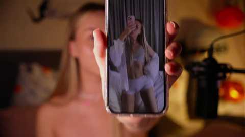 Video of a blonde woman in lingerie, reflected in a smartphone, held by a hand. Background blurred, featuring a camera and warm, dimly lit room with a hint of red.