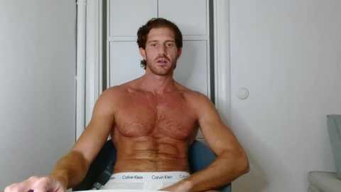 Video of a shirtless, muscular, white man with short brown hair, sitting in Calvin Klein underwear in a dimly lit room with white walls and a door.