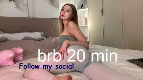 Video of a slim, pale-skinned woman with long blonde hair, wearing a strapless green top and tight white leggings, sitting seductively on a bed. Background features a plush, gray headboard, fluffy pink pillow, and a soft white blanket. Text overlay reads \"bbr 20 min Follow my social.\