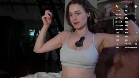 Video of a young white woman with curly hair, wearing a light gray sports bra and a necklace, dancing in a dimly lit room.