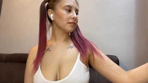 Video of a young woman with medium skin tone and long, straight, maroon-colored hair in a high ponytail. She wears white headphones, a white tank top, and has small, black tats on her collarbones. She sits on a brown sofa against a beige wall.