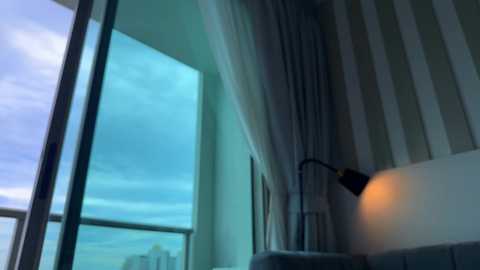Video of a modern hotel room with large windows showing a cityscape. A black floor lamp and striped curtains are visible.