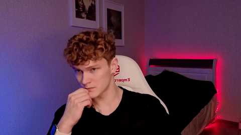 Video of a young, curly-haired man in a black shirt, sitting on a white gaming chair with a red LED strip behind him.