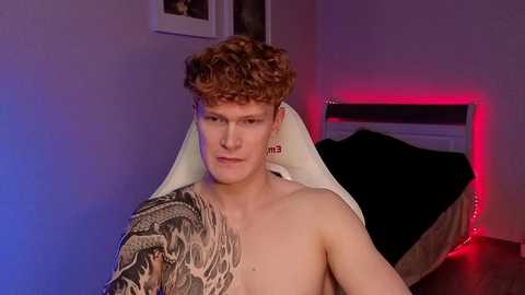 Video of a shirtless young man with curly red hair and a large, intricate tattoo on his right shoulder. He sits on a bed with black and white bedding, under dim purple and red lighting.