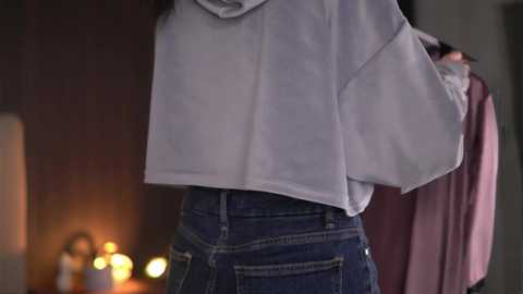 Video of a person wearing a white, oversized hoodie and high-waisted, dark blue jeans, standing indoors near a dimly lit, warm-colored wall.