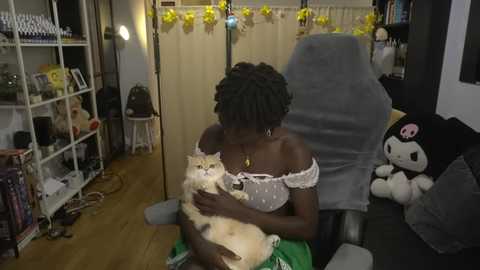 Video of a dark-skinned woman with short, curly hair, wearing a white off-shoulder top, holding a fluffy white cat. Background features a beige curtain, white shelves, stuffed toys, and a gaming chair.